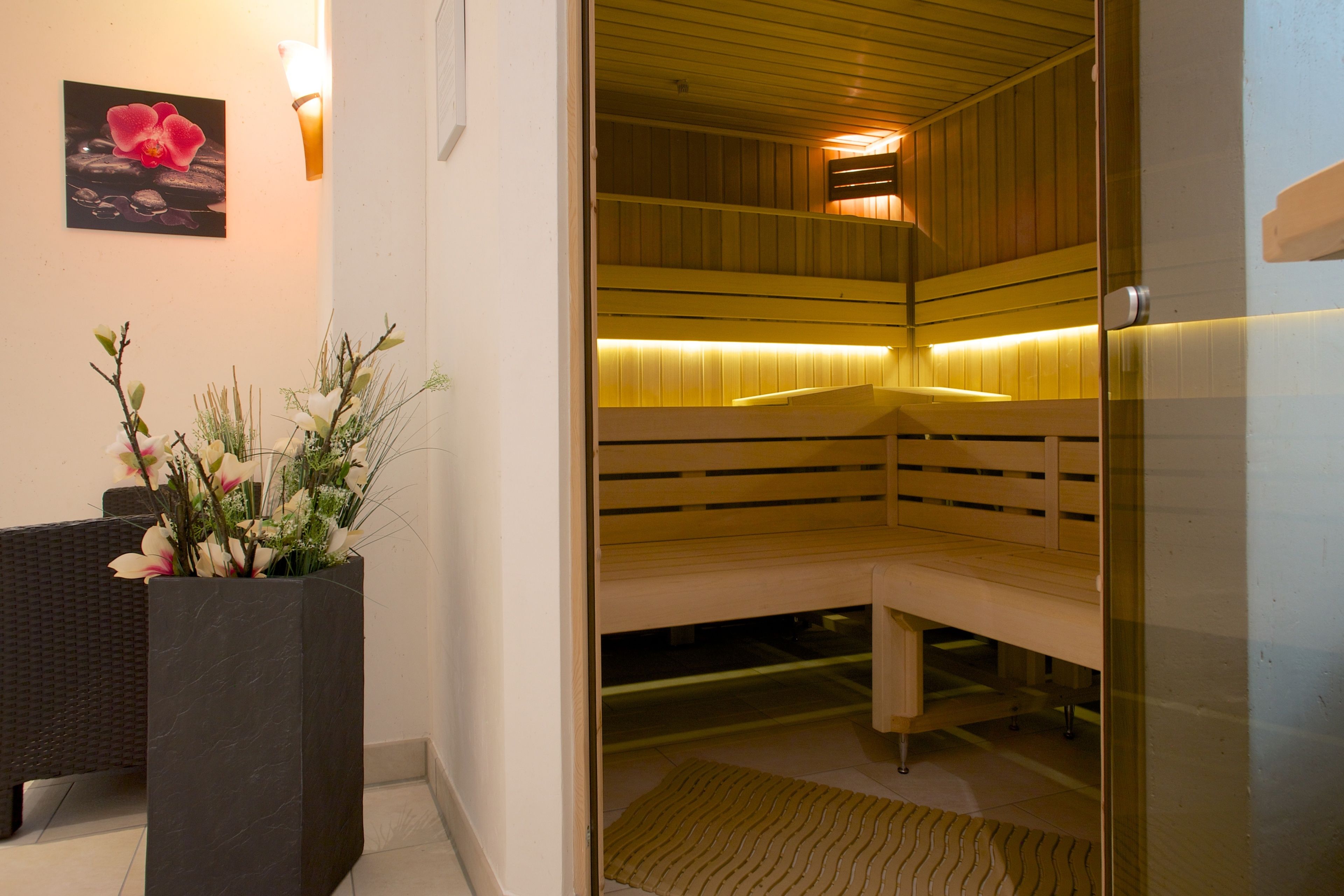 A sauna with wooden benches and a glass door, a dark marble planter with flowers, and a wall-mounted picture of a flower.
