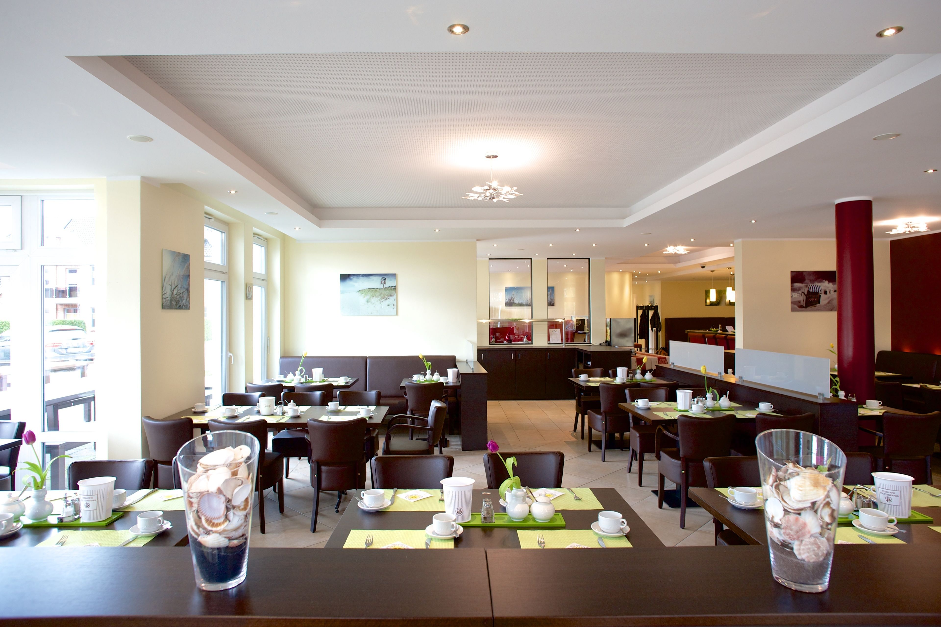 A spacious dining area with multiple tables and chairs, a kitchen in the background, and a view of the outside through large windows.