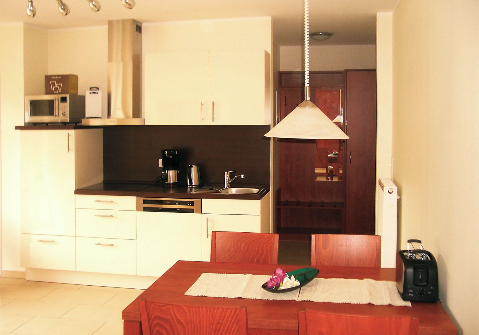 A compact kitchen with white cabinets, a microwave, a coffee maker, and a sink. There's a dining table with a placemat and a green bowl.