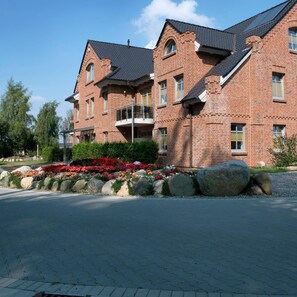 A multi-story brick building with a symmetrical facade, large windows, and a landscaped garden with colorful flowers and rocks. - Aparthotel Museumshof (Fehmarn)