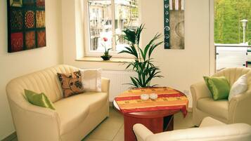 A living room with two cream-colored sofas, a small round table with a colorful tablecloth, a potted plant, and a window with a view of buildings and greenery.
