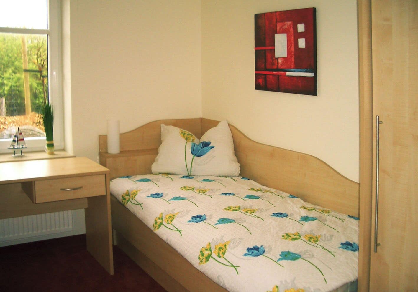 A bedroom with a wooden bed, a bedside table, a window with a view of greenery, and a red abstract painting on the wall.