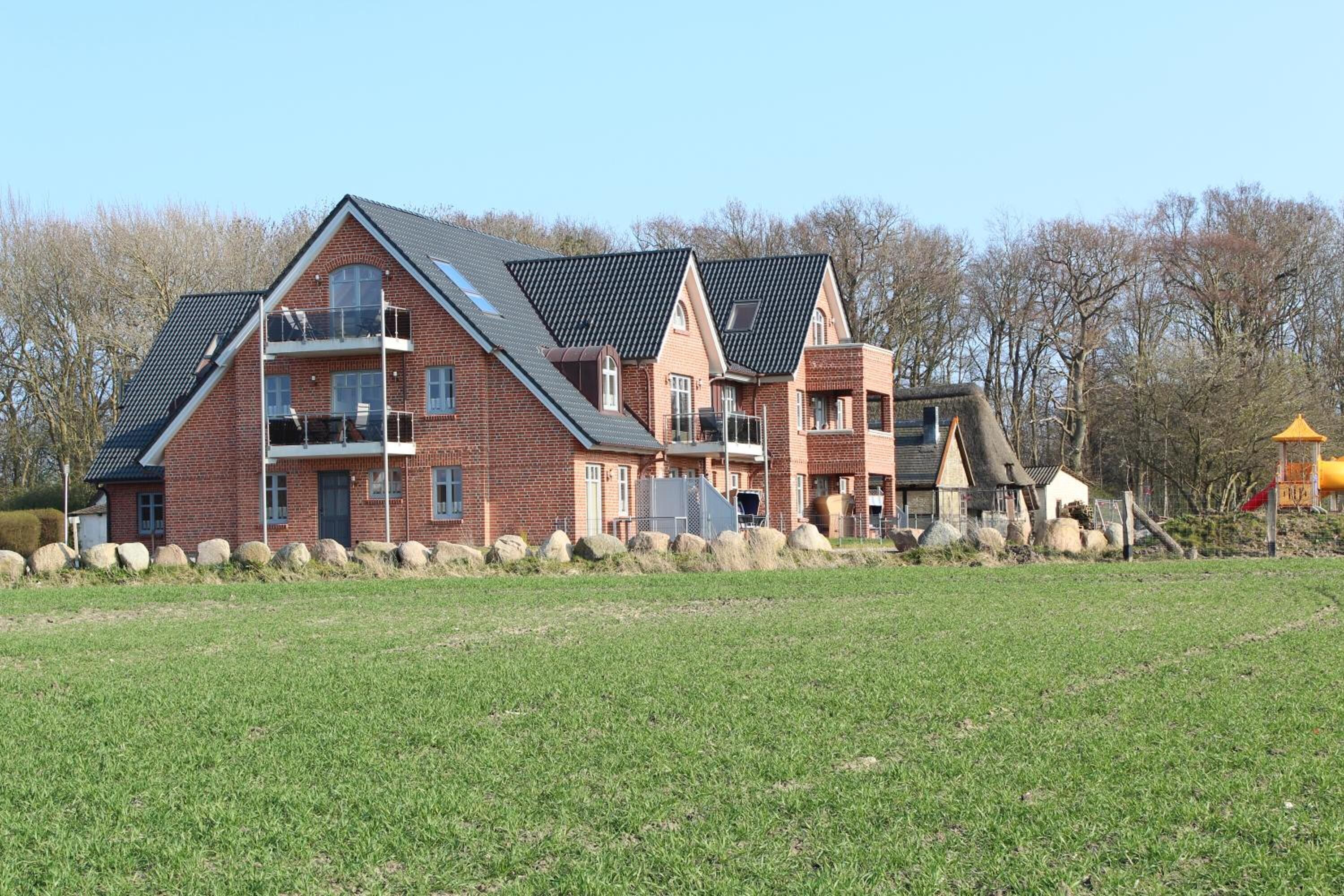 Une grande maison en briques, à plusieurs étages et dotée d’un toit en pente, entourée d’un champ verdoyant et d’arbres.