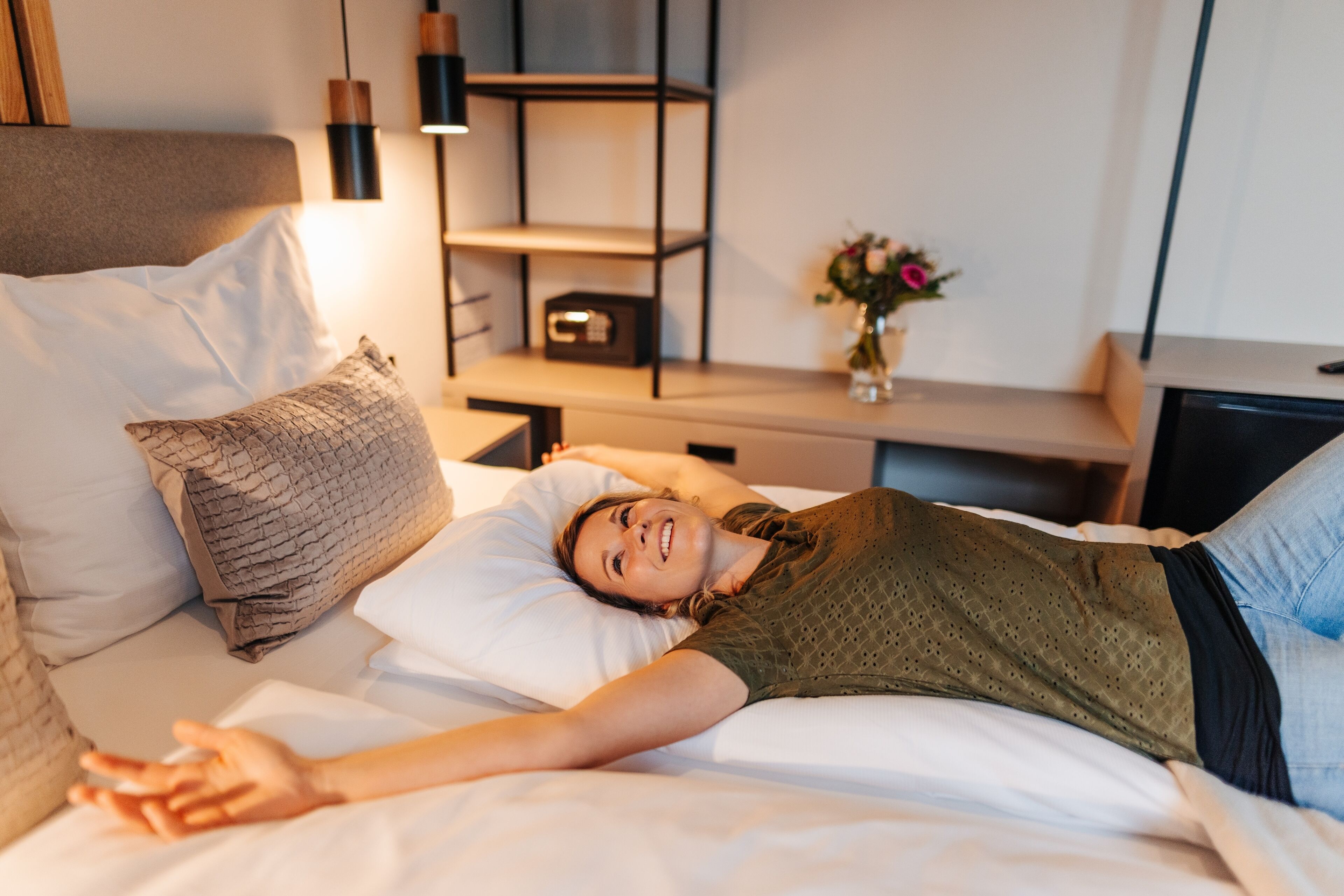 A person lying on a bed with a headboard, a bedside table with a lamp, and a small shelf with a vase of flowers.