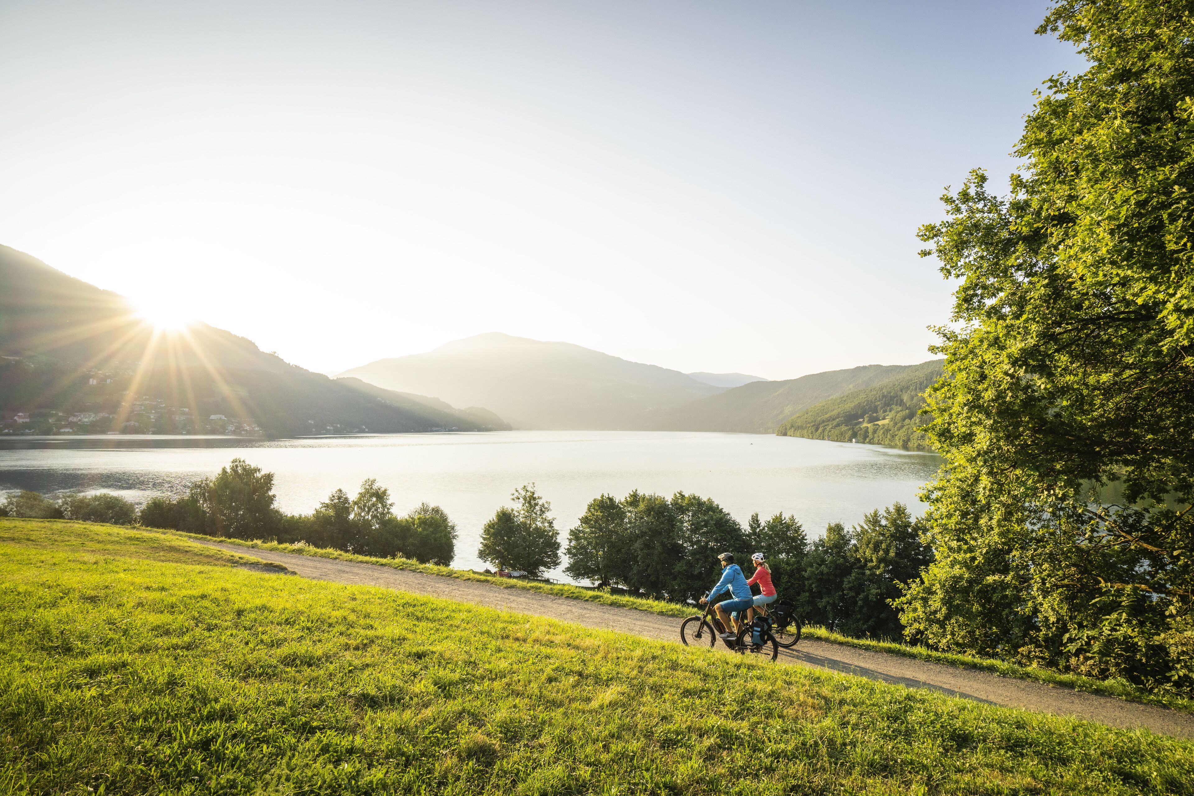 A scenic view of a lake with mountains in the background and a path with cyclists.