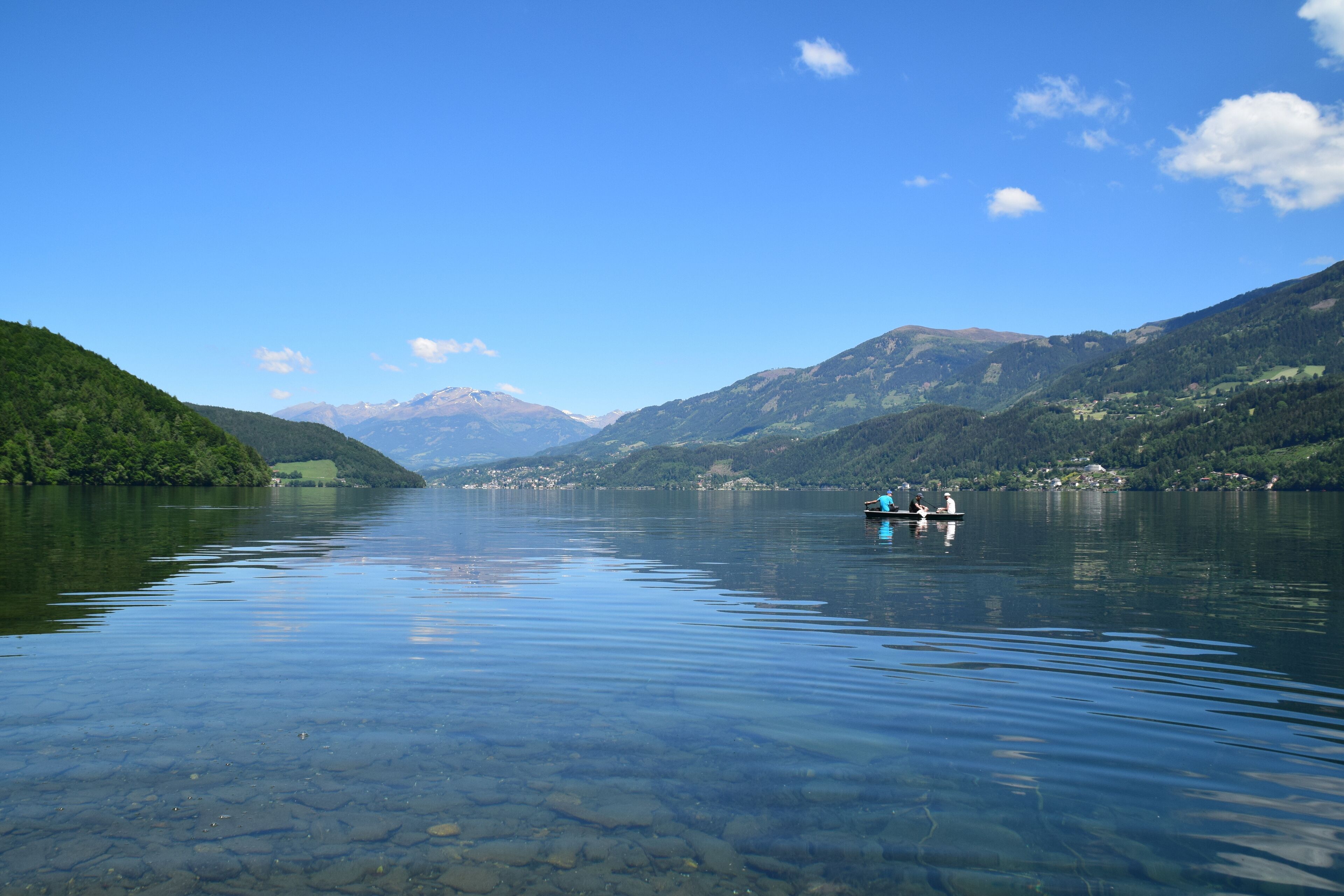A calm lake with clear water, surrounded by green hills and mountains in the distance.