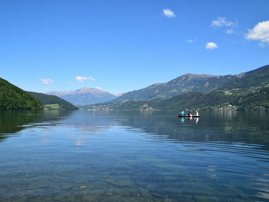 A calm lake with clear water, surrounded by green hills and mountains in the distance.