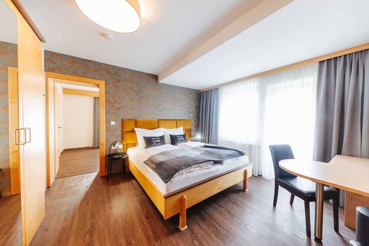 A hotel room with a wooden bed, a desk, and a chair. There is a door leading to another room and a window with curtains.