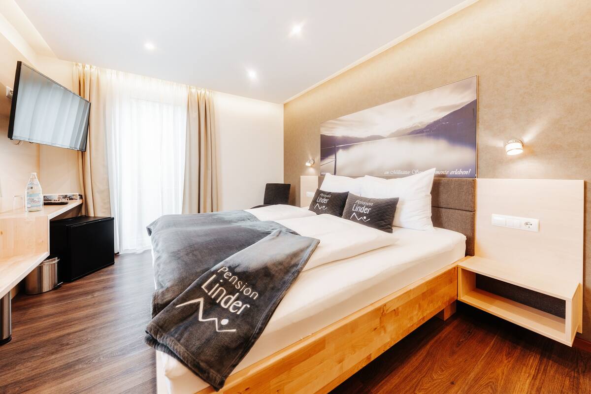 A modern hotel room with a large bed, a TV mounted on the wall, a desk with a lamp, and a view of the mountains through the window.