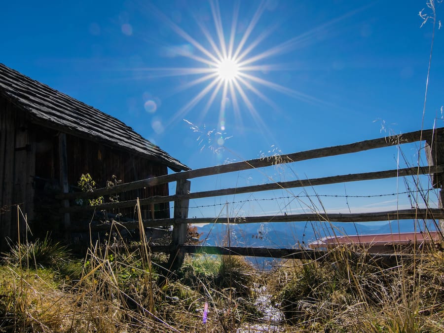 A rustic wooden barn with a metal fence in front, set against a clear blue sky with the sun shining brightly.