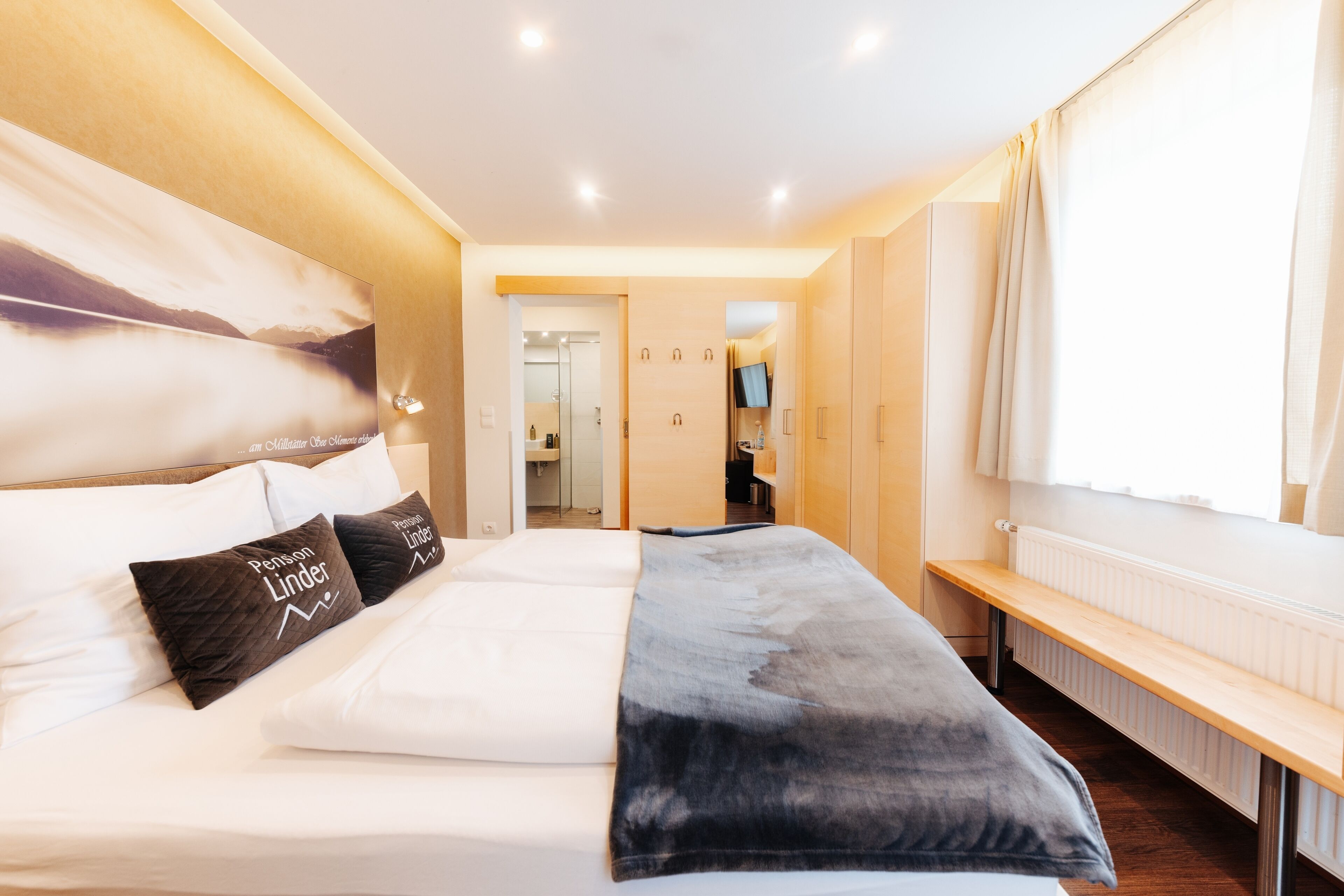 Un cuarto de hotel con una cama grande, un banco, un televisor y vista al pasillo.