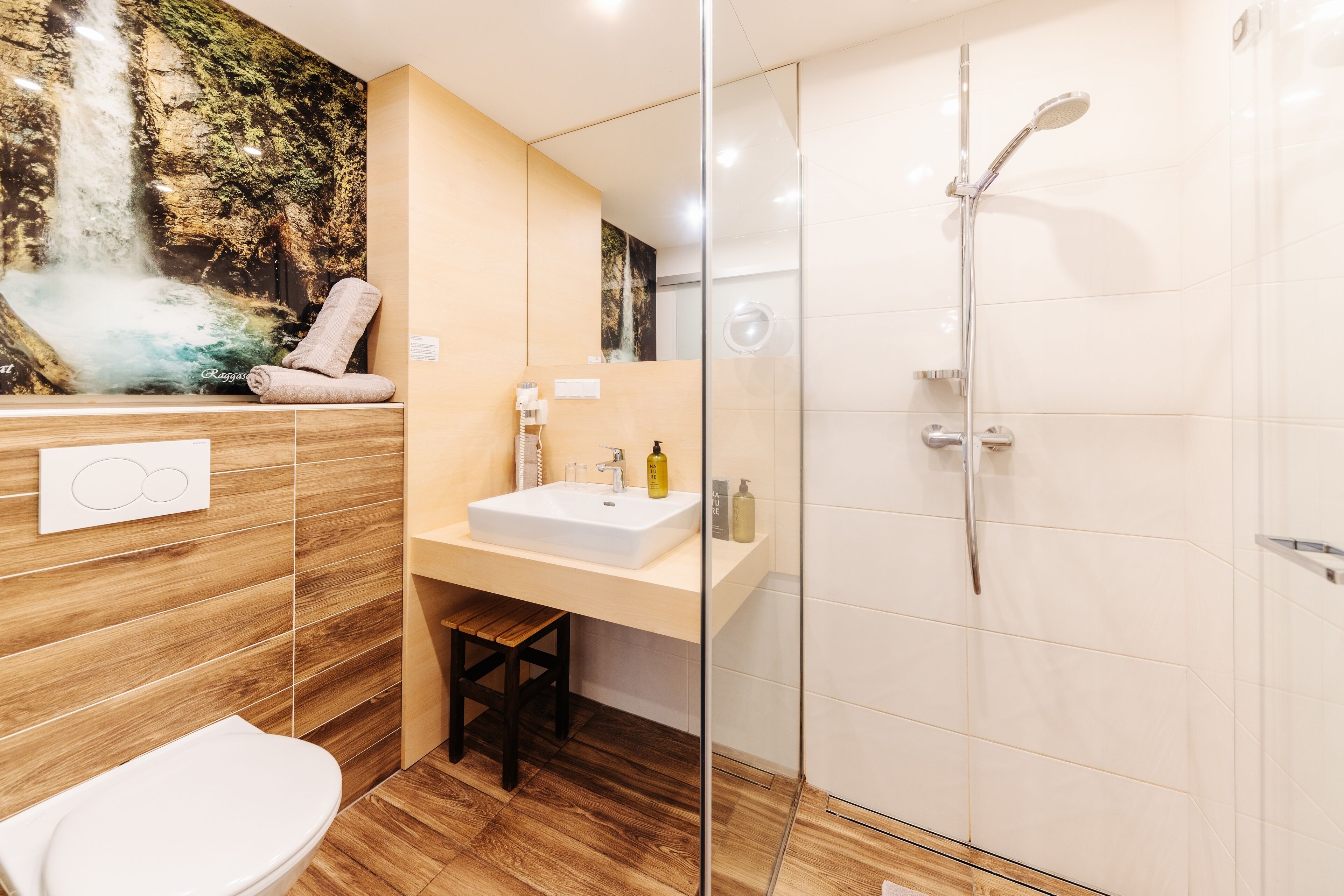 A modern bathroom with a wooden vanity, a glass shower enclosure, and a waterfall mural on the wall.