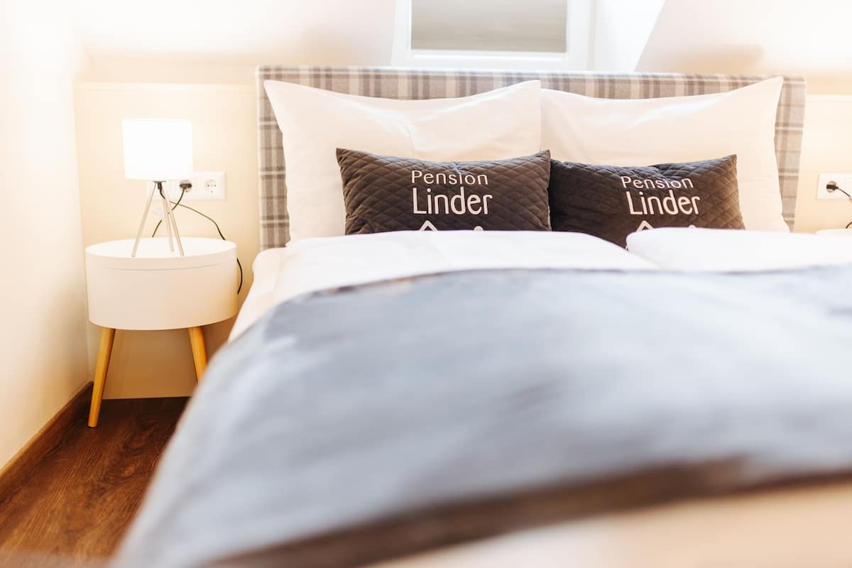 A neatly made bed with two pillows labeled "Pension Linder" and a bedside lamp on a white nightstand.