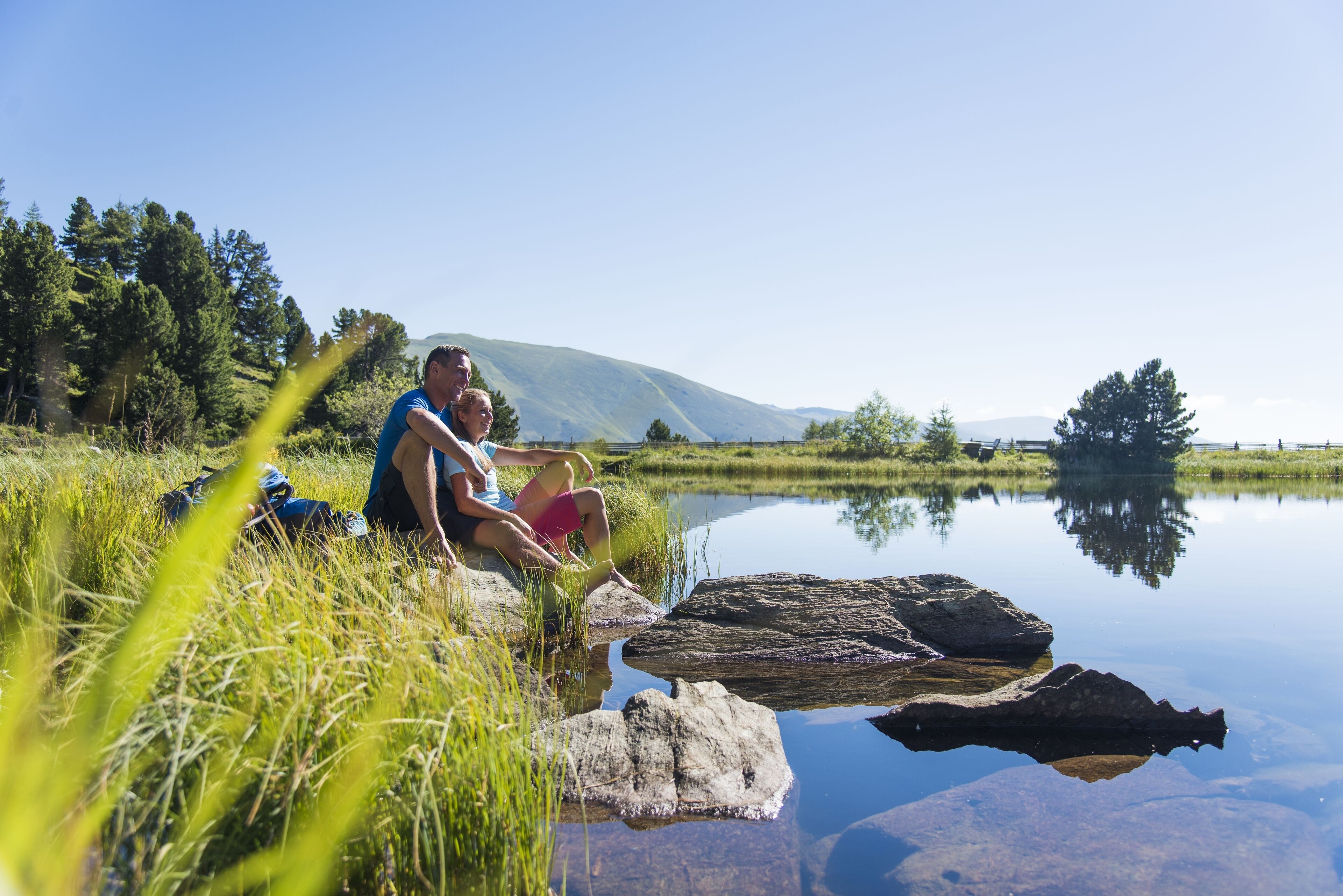 A couple sitting by a calm lake with mountains in the background.