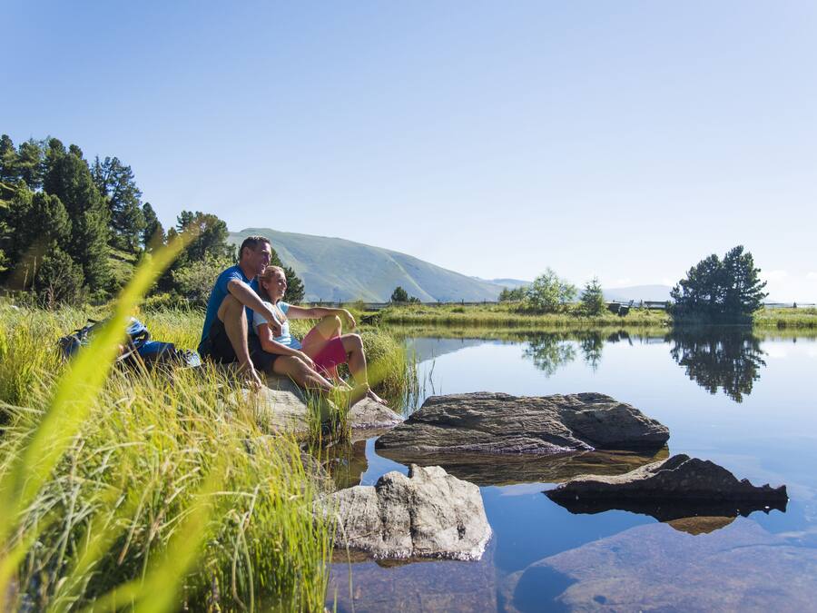 A couple sitting by a calm lake with mountains in the background.
