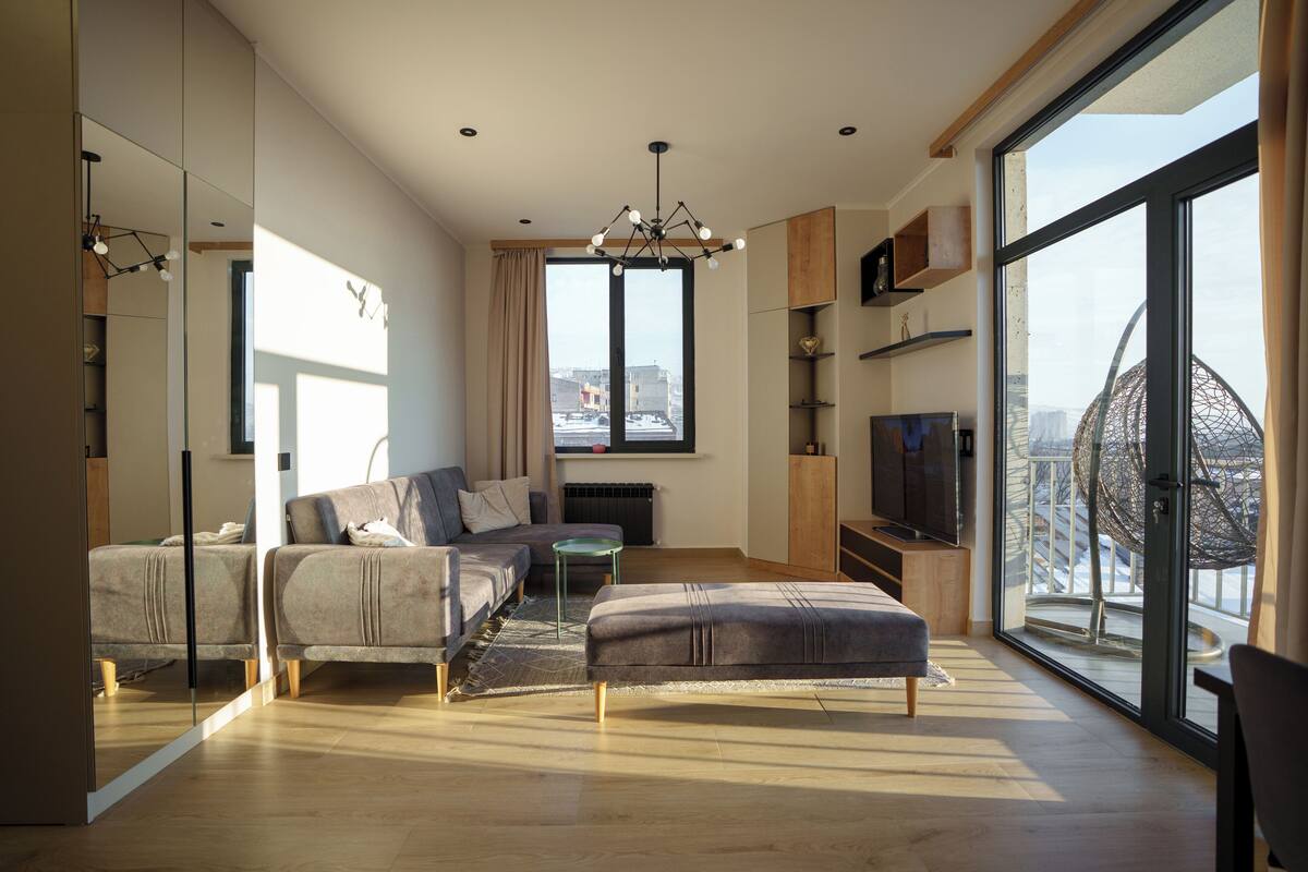 A modern living room with a sofa, ottoman, and TV, large windows with a city view, and a chandelier.