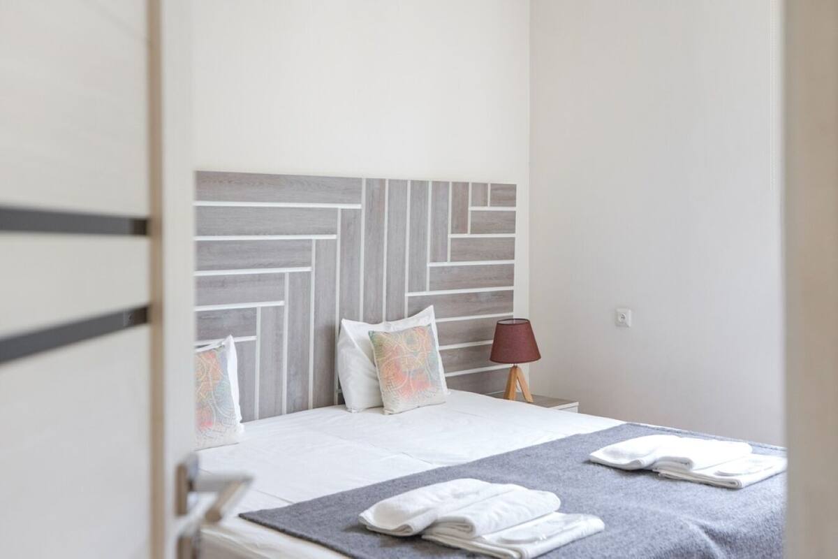 A neatly made bed with a grey and white headboard, a bedside lamp, and folded towels on the floor.