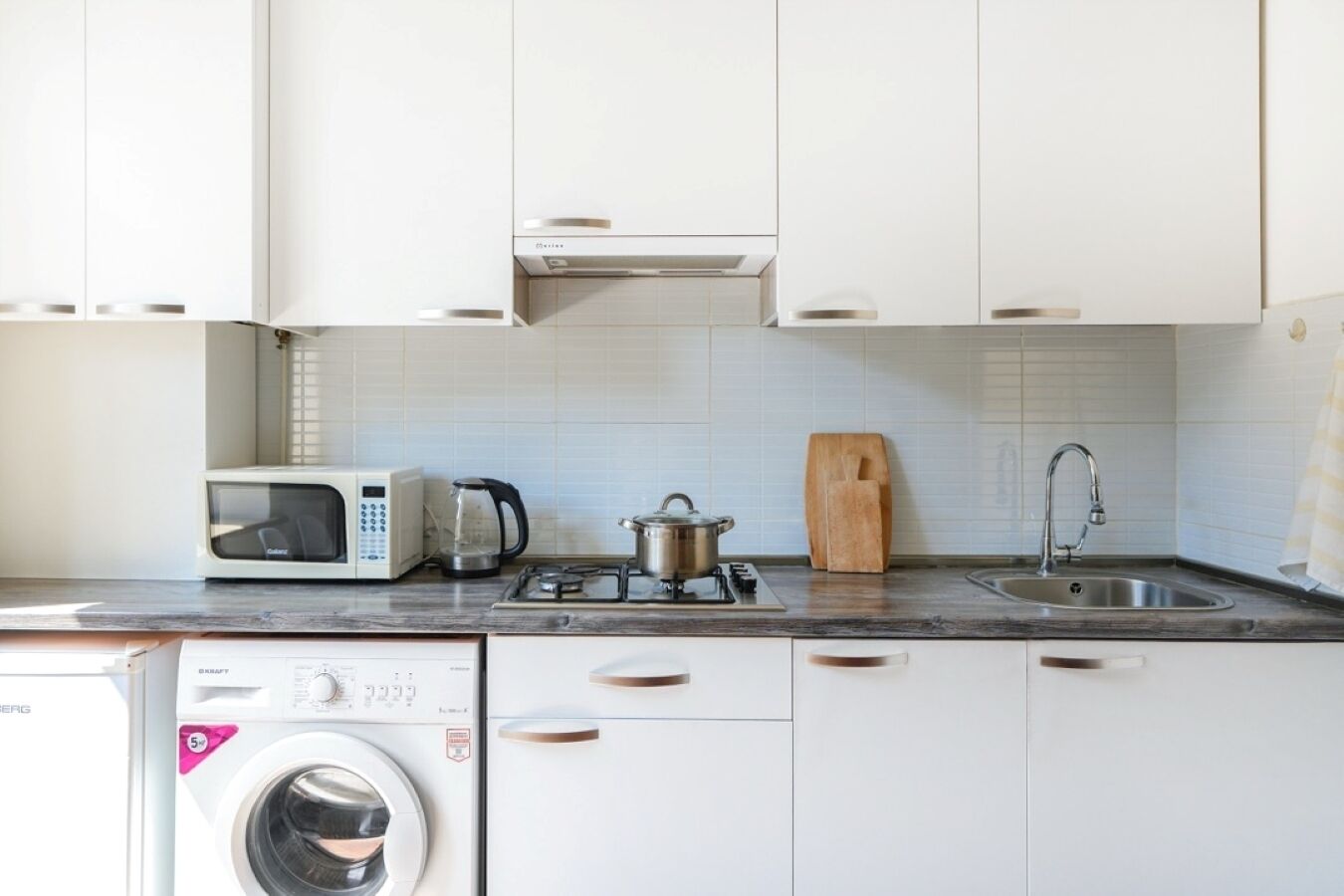 Cocina compacta con armarios blancos, microondas, estufa a gas, lavamanos y lavadora.