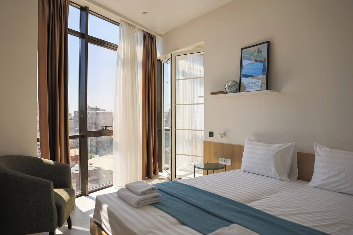 A hotel room with a bed, a chair, a small table, a window with curtains, and a framed picture on the wall.