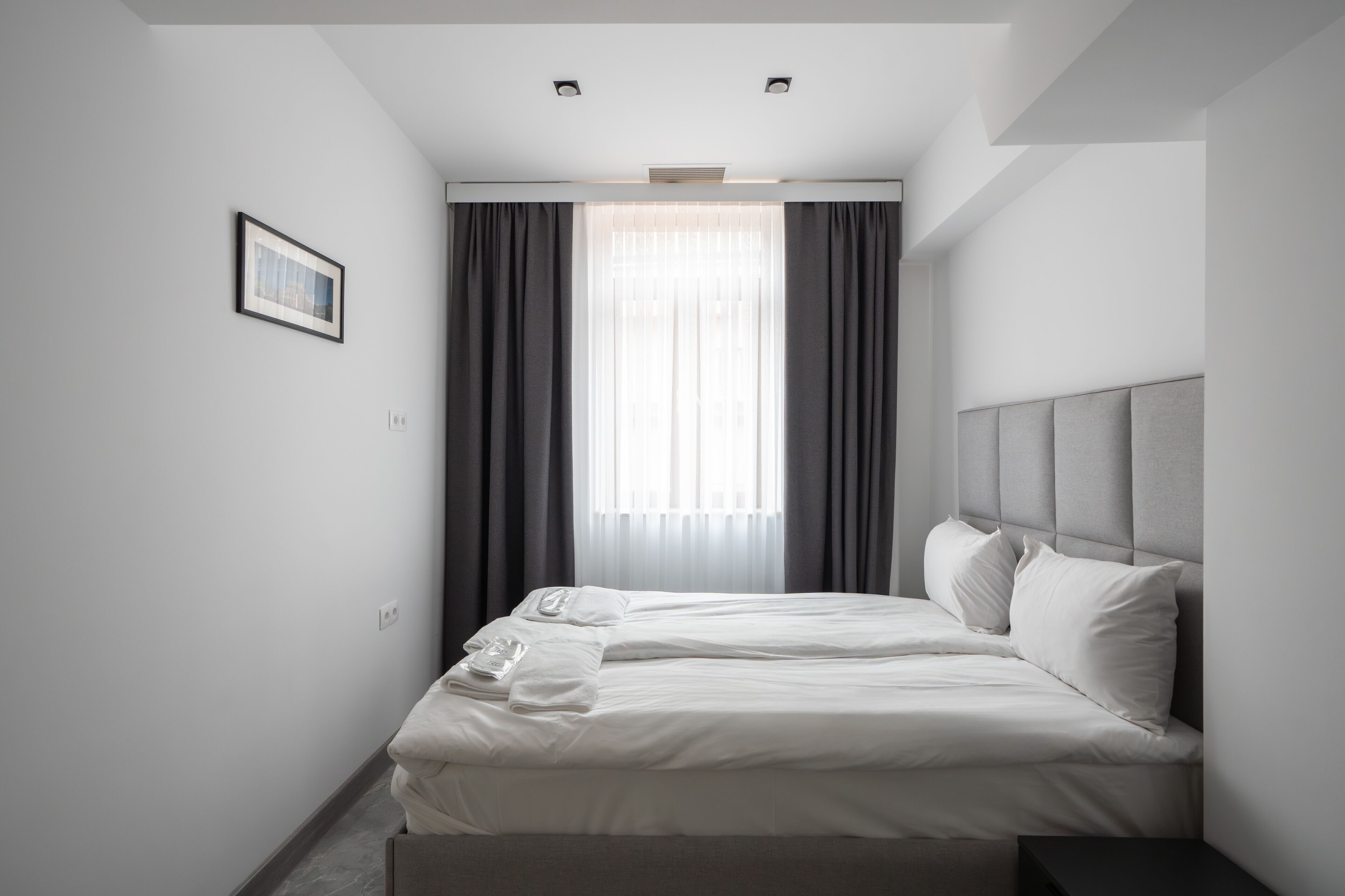 Ein modernes Hotelzimmer mit einem ordentlich bezogenen Bett, einem großen Fenster mit Vorhängen und einem gerahmten Bild an der Wand.