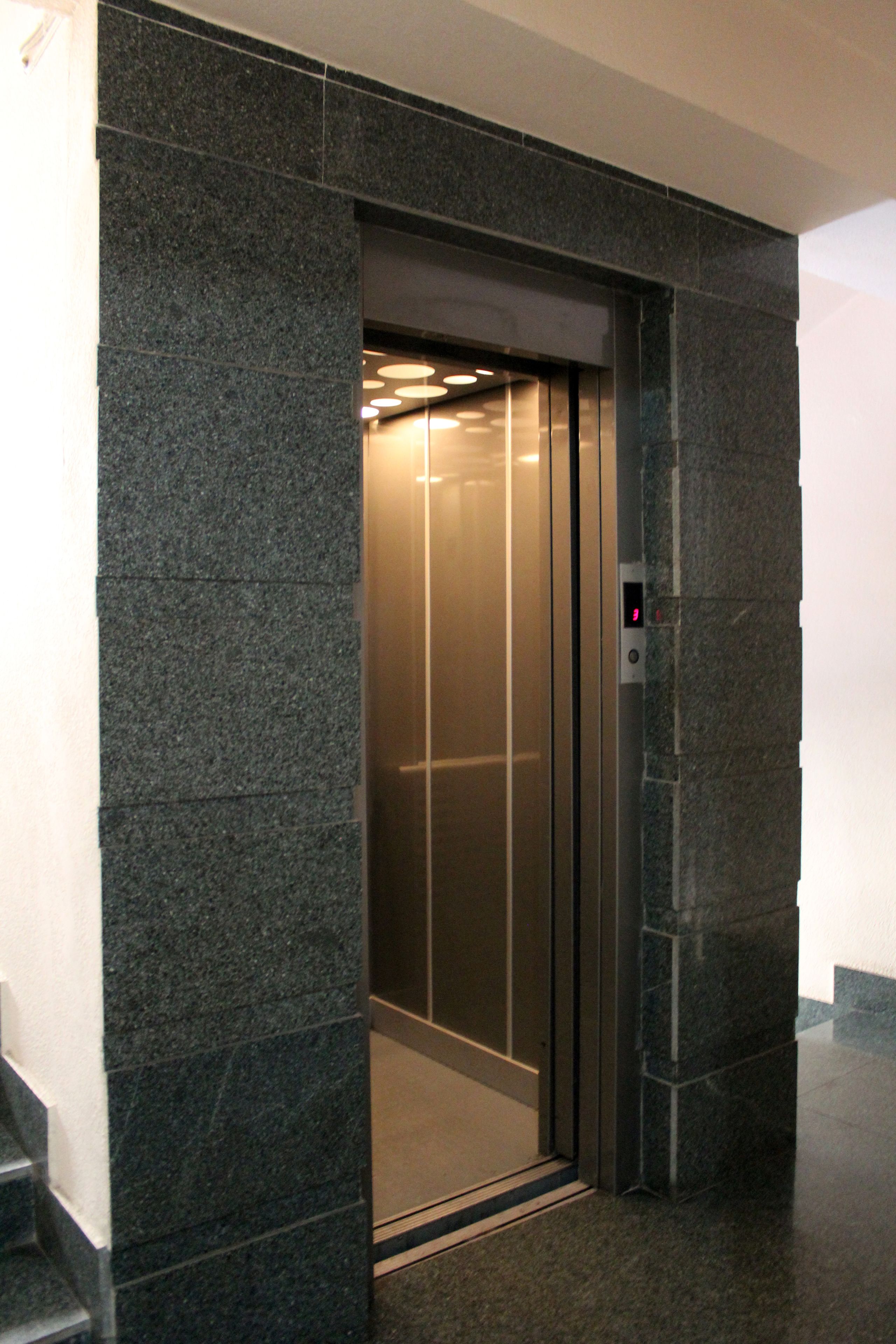 En elevator med digitalt display, omgivet af mørke stenvægge og et flisebelagt gulv.