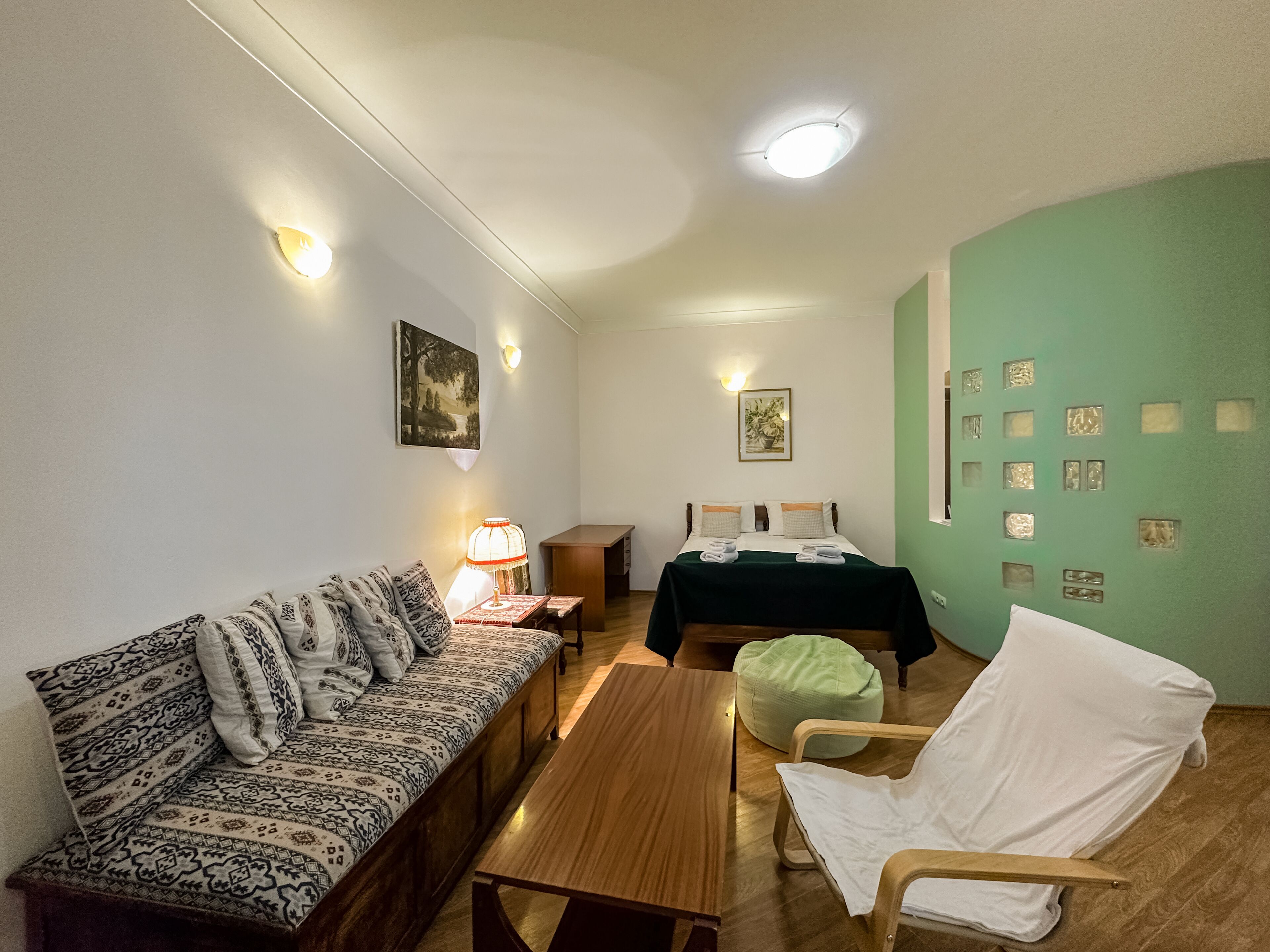 A cozy room with a patterned sofa, a wooden table, a green bed, and a white chair.