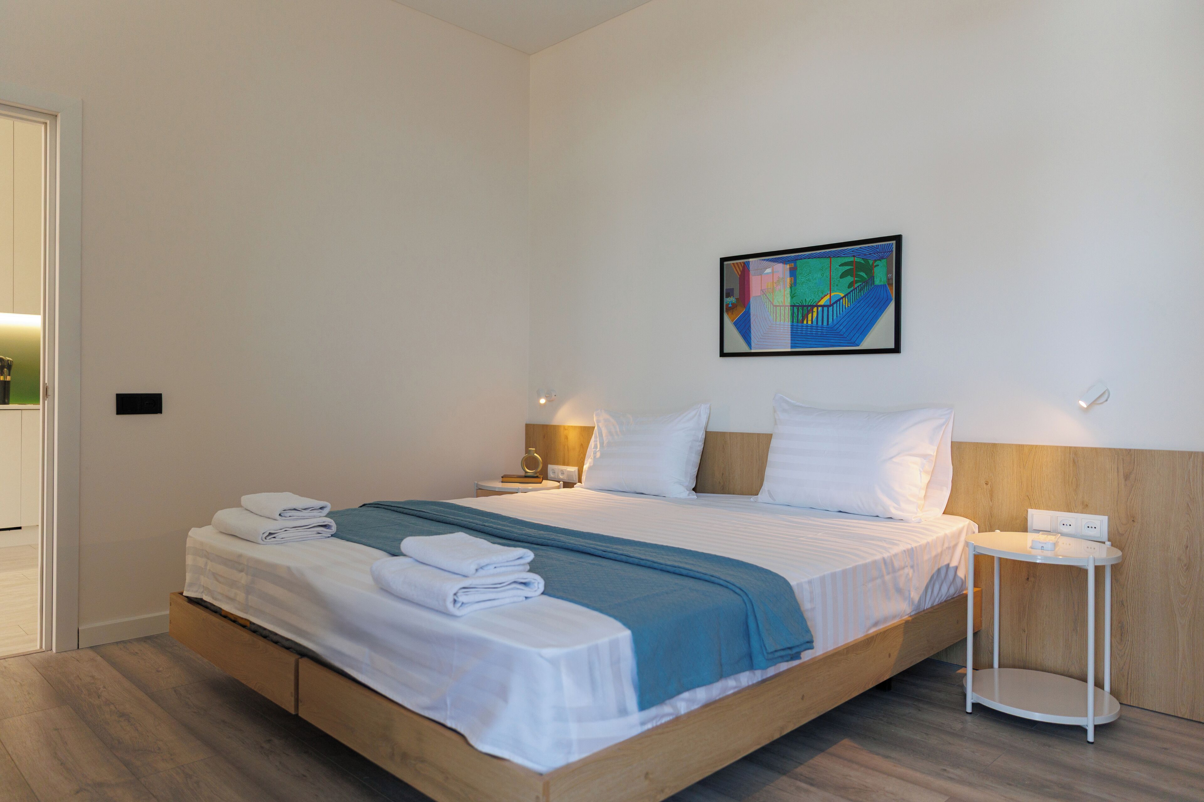 Un lit bien fait, avec une couverture bleue, des oreillers blancs et des serviettes pliées. Il y a une petite table de chevet avec une lampe, et un tableau accroché au mur au-dessus du lit.