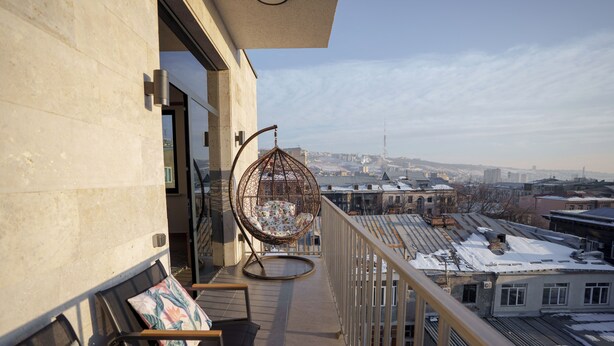 A balcony with a hanging chair and a cushioned chair, overlooking a cityscape with snow-covered buildings. - RIS Central Apartments Yerevan (Yerevan)