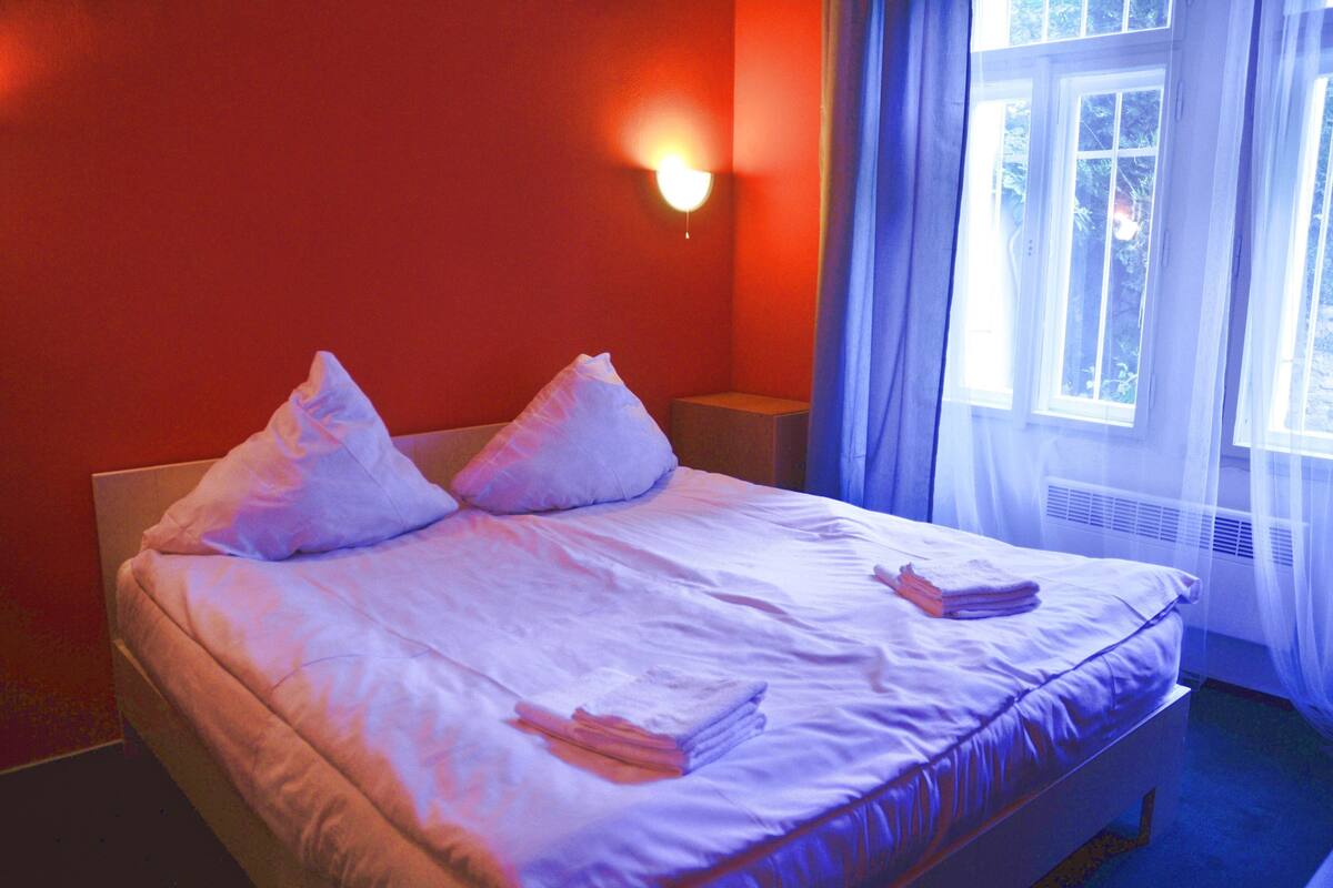 Una cama con sábanas y almohadas blancas, una mesita de noche y una ventana con cortinas.