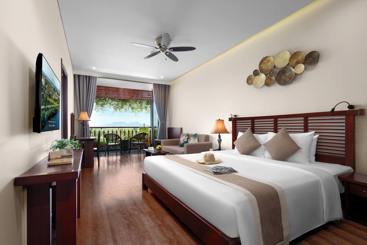 A hotel room with a bed, bedside tables, a desk, a chair, a ceiling fan, and a view of the outdoors.