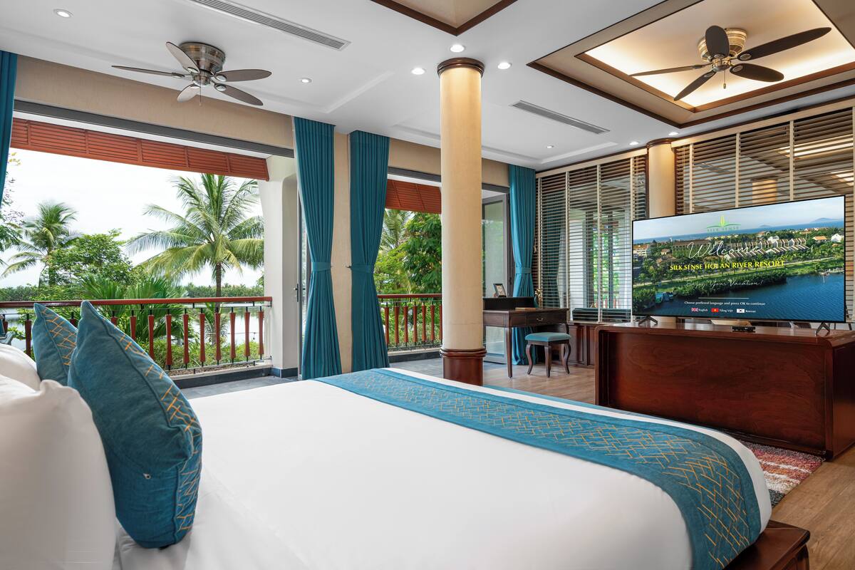 A hotel room with a large bed, a desk, a television, and a balcony with a view of palm trees.