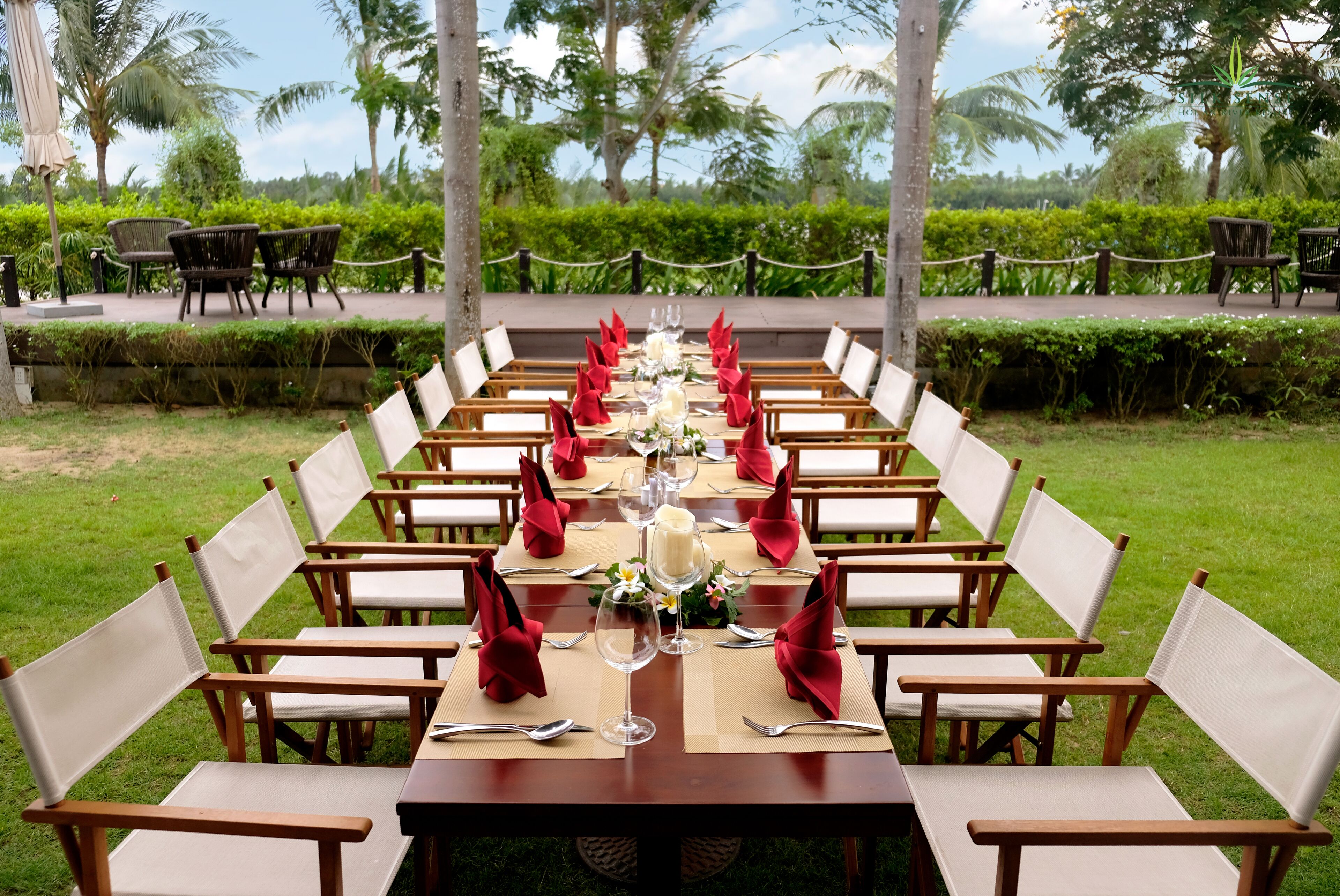A long dining table set outdoors with white chairs and red napkins.