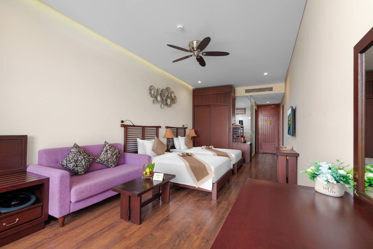 A hotel room with a purple sofa, two beds, a ceiling fan, wooden furniture, and a small table with a plant.