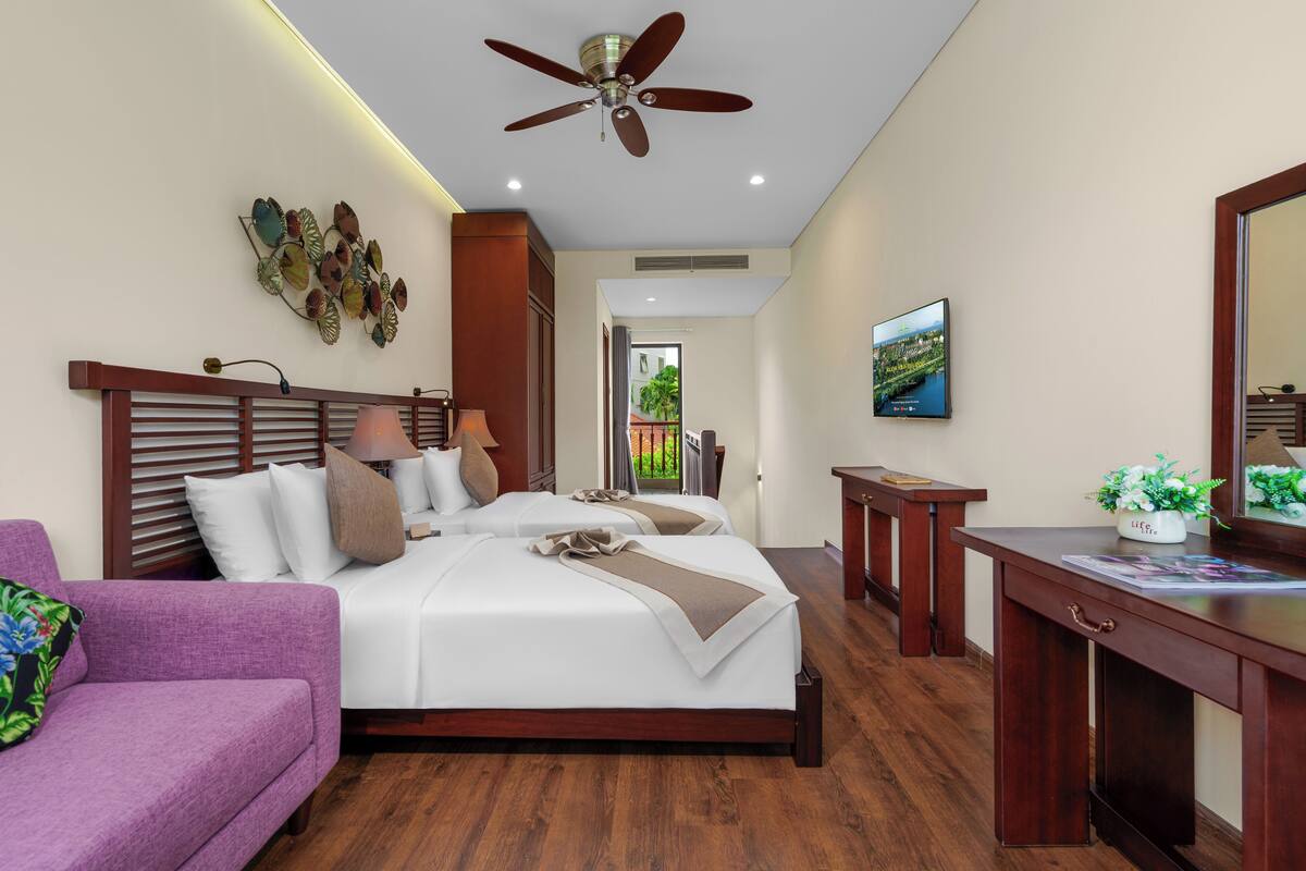 A hotel room with a bed, a desk, a purple sofa, a ceiling fan, and a wall-mounted TV.