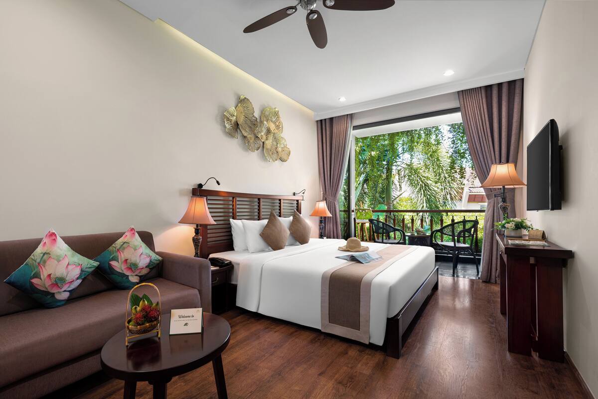 A hotel room with a bed, a sofa, a coffee table, a TV, and a balcony with a view of greenery.
