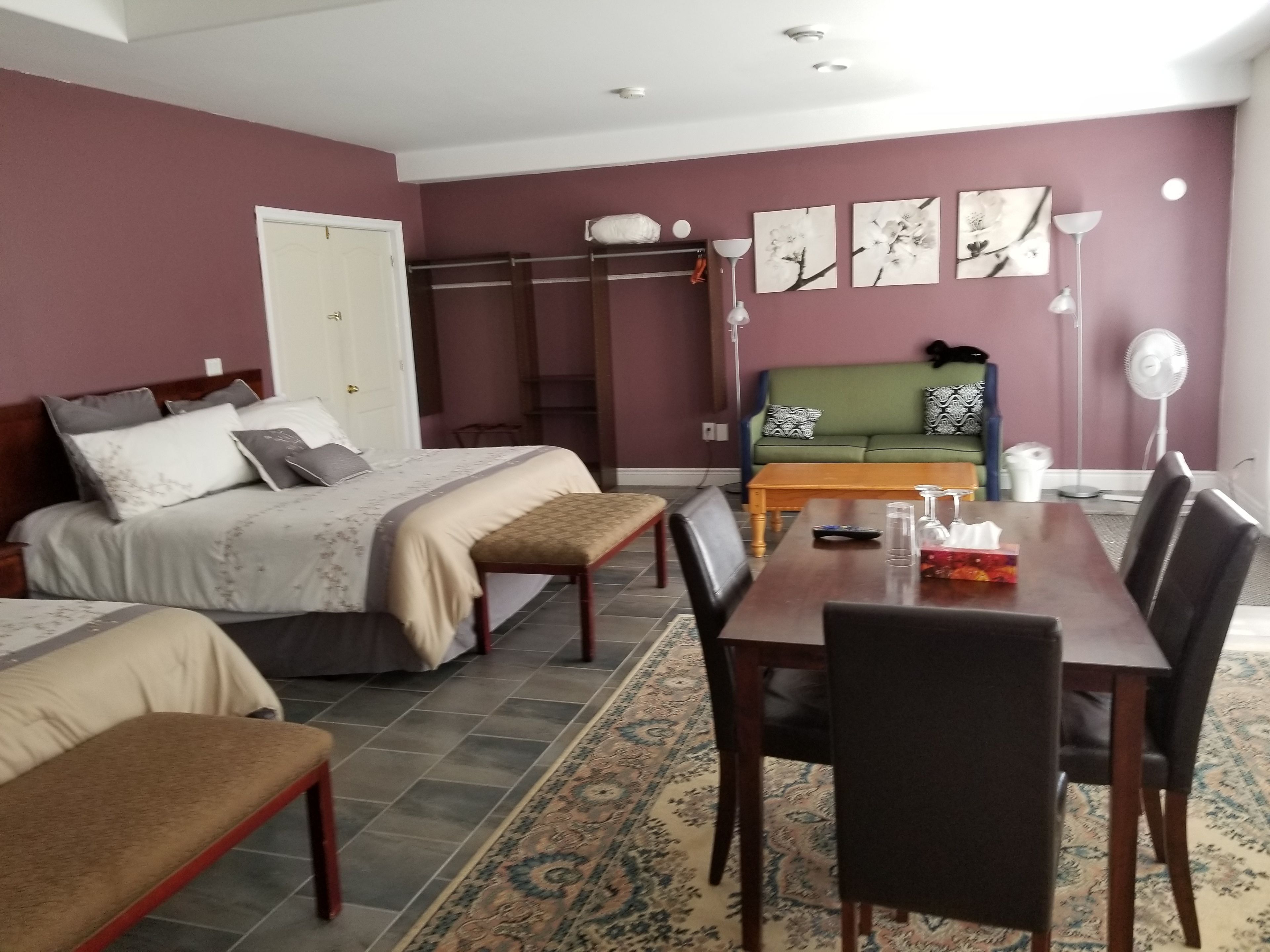 A bedroom with a bed, bedside tables, a bench, a dining table with chairs, a sofa, and a fan.
