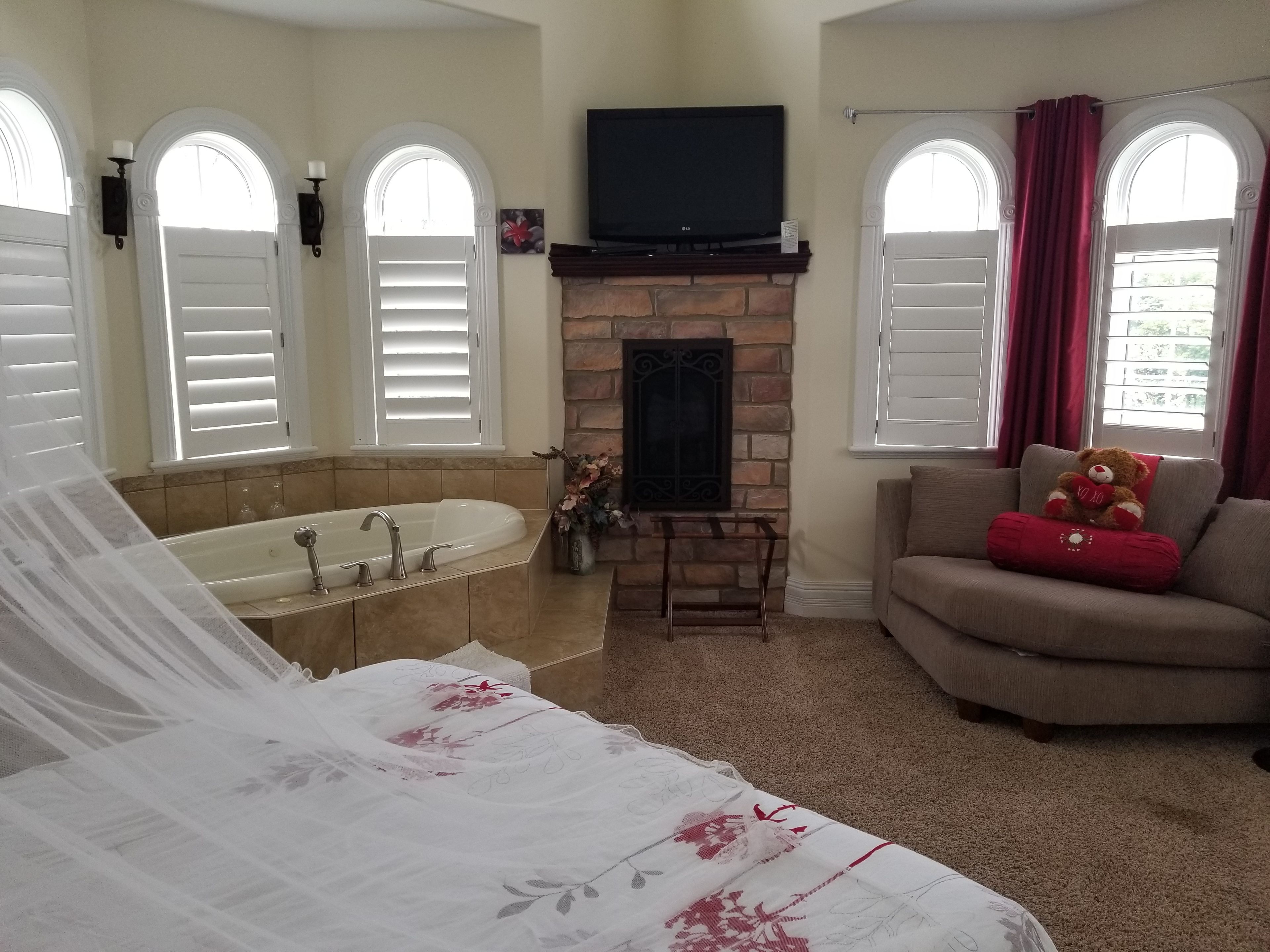 A bedroom with a large bed, a bathroom with a bathtub, a fireplace, and a living area with a sofa and a teddy bear.