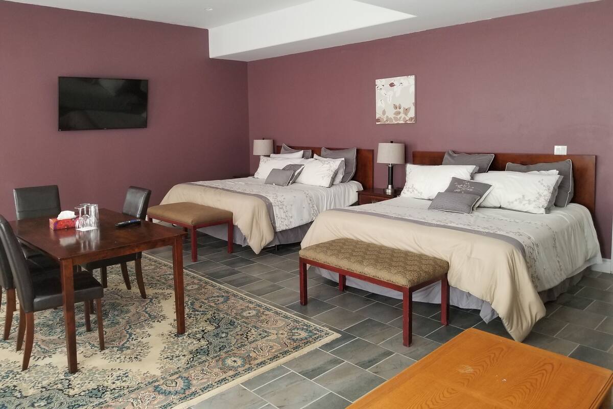 A hotel room with two beds, a desk, chairs, a TV, and a large window.