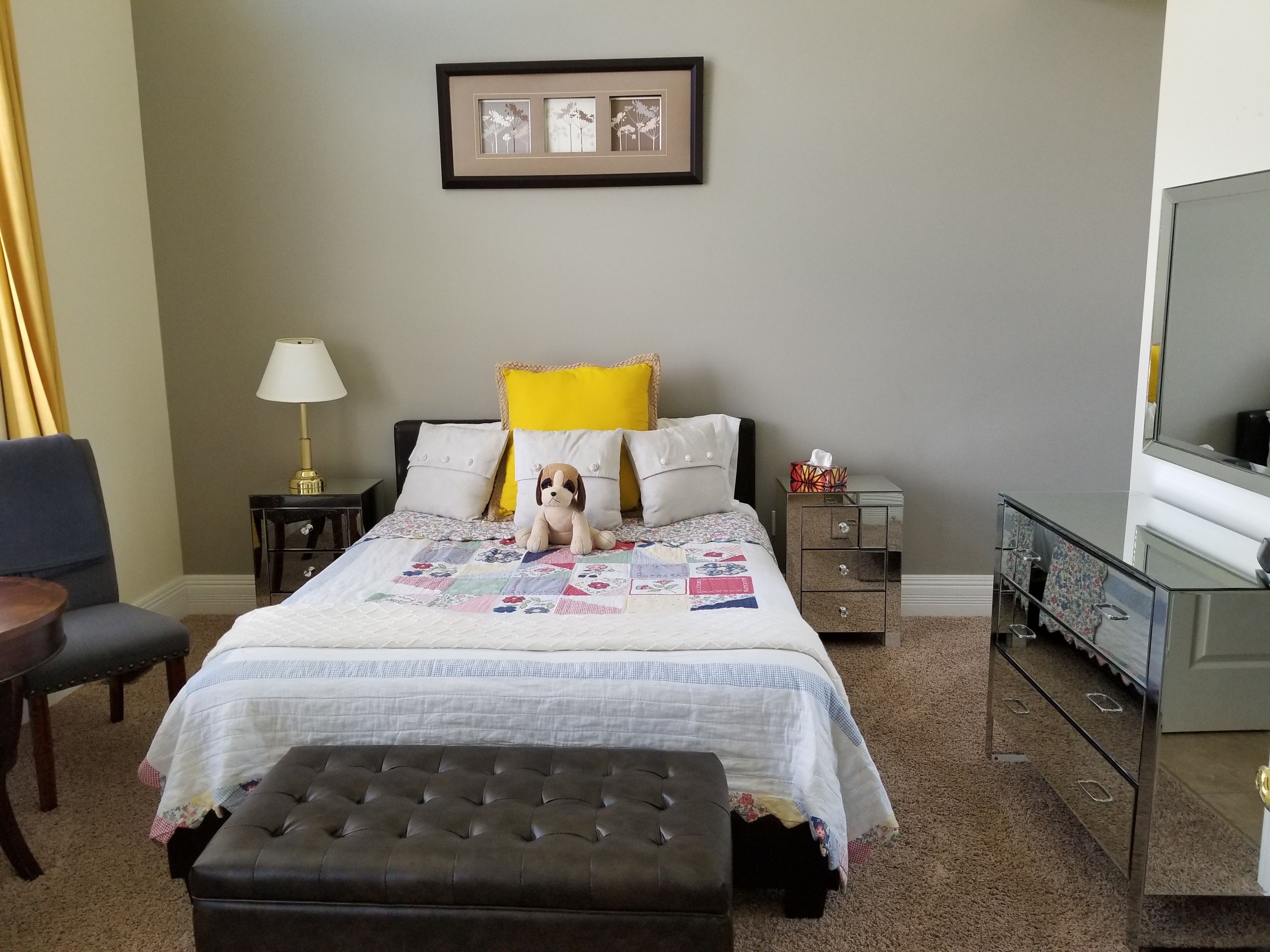 A neatly arranged hotel room with a bed, a small table, a chair, a bedside lamp, a bedside table with a lamp, a dresser, and a mirror.