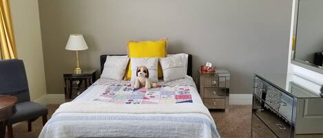 A neatly arranged hotel room with a bed, a small table, a chair, a bedside lamp, a bedside table with a lamp, a dresser, and a mirror.