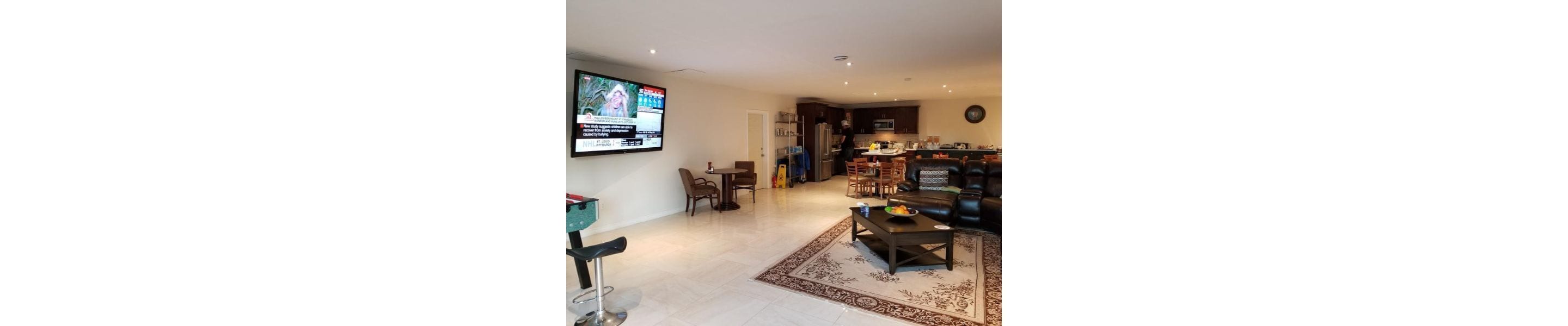 A modern living room with a flat-screen TV mounted on the wall, a dining area with a table and chairs, and a kitchen area with a refrigerator and microwave.