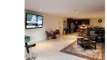 A modern living room with a flat-screen TV mounted on the wall, a dining area with a table and chairs, and a kitchen area with a refrigerator and microwave.