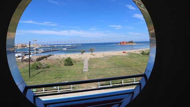 Vista a través de un ventanal circular de un puerto deportivo con barcos, una playa y un edificio rojo distante.