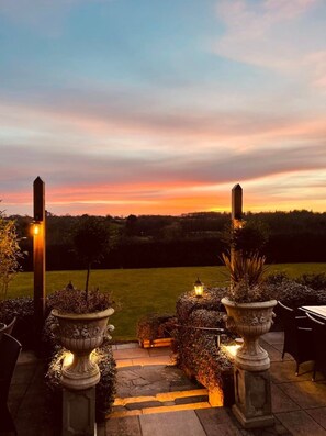 An outdoor patio area with stone columns, potted plants, and a view of a sunset over a grassy field.