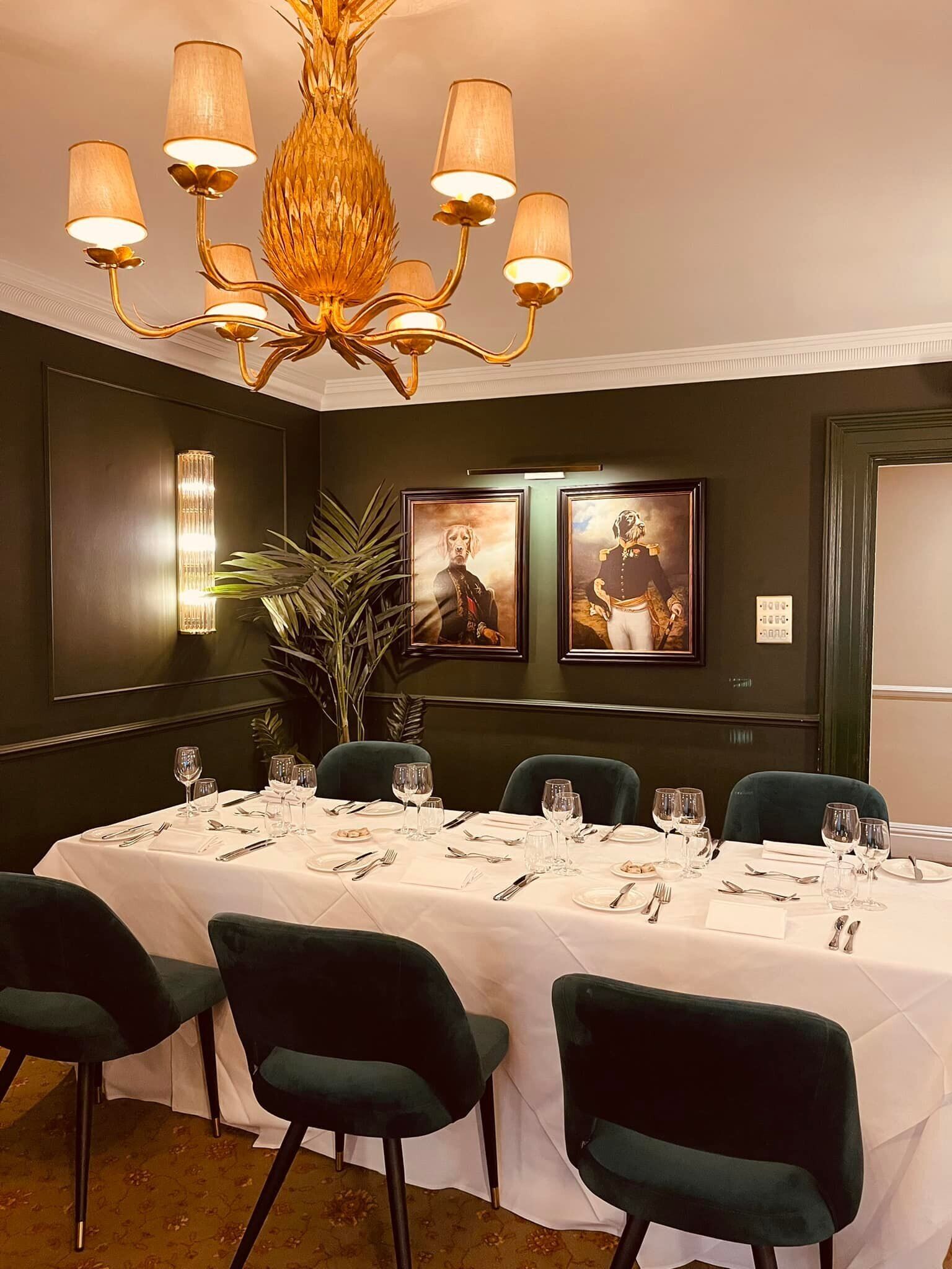 Comedor con una larga mesa dispuesta para seis comensales, sillas tapizadas en verde, una araña, retratos enmarcados en la pared y una planta en maceta.