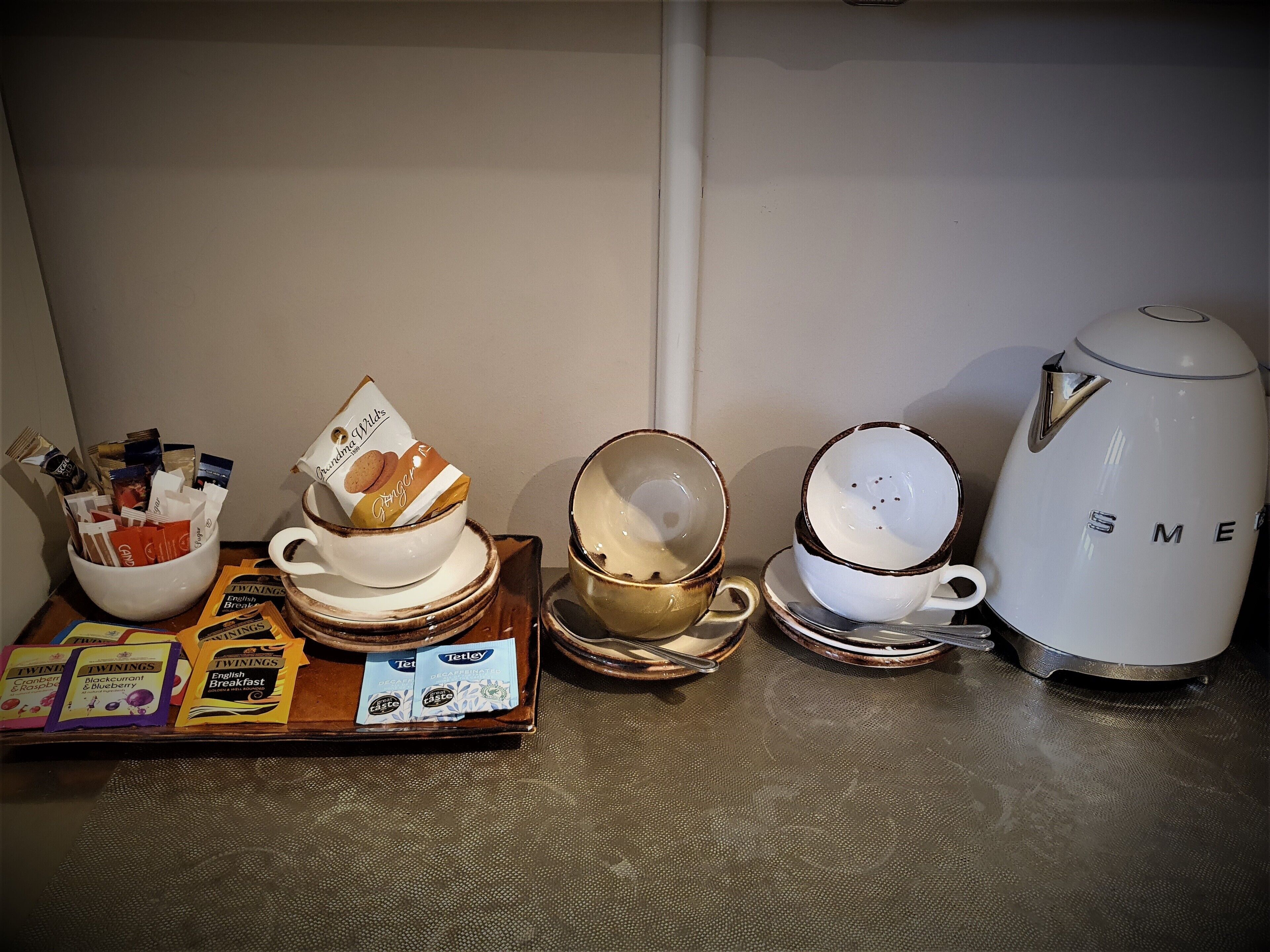 A kitchen counter with various items including a white kettle, stacked bowls, a tray with breakfast cereal packets, and a bowl of cookies.