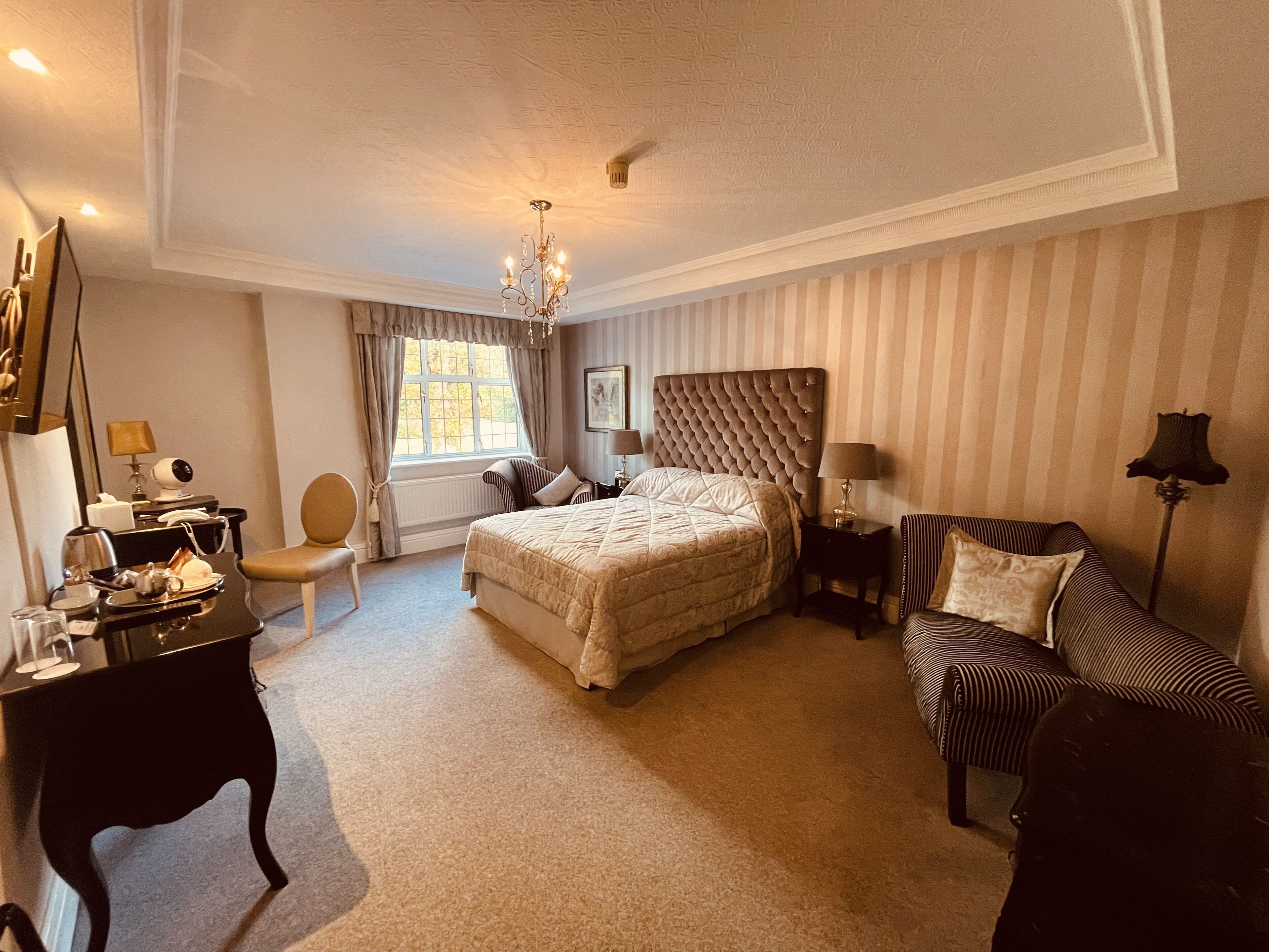 Ein Hotelzimmer mit einem großen Bett, einer Sitzecke inklusive Sofa und Sessel, einem Schreibtisch mit Telefon und Lampe sowie einem Fenster mit Vorhängen.