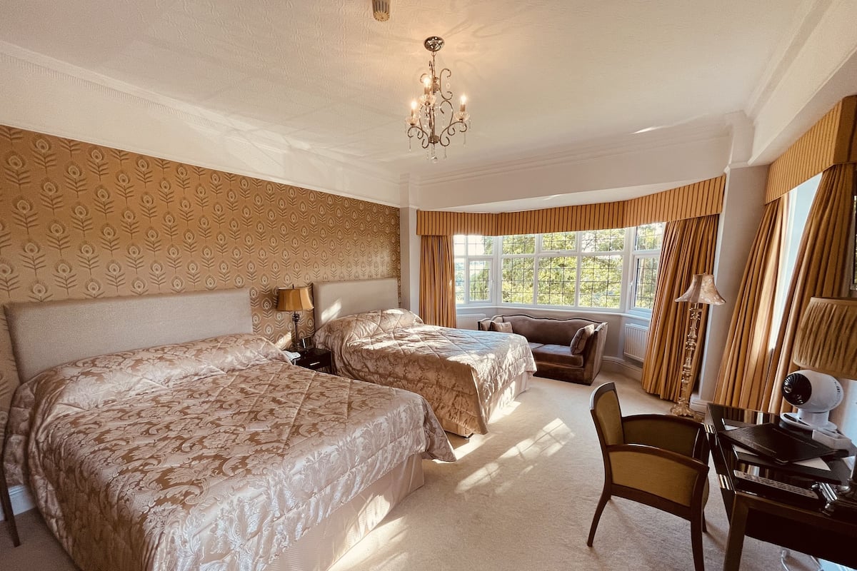 Ett hotellrum med två sängar, ett skrivbord, en stol, en kristallkrona och stora fönster med gardiner.