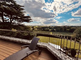Una terraza con una silla de descanso, baranda de piedra y vista a un paisaje verde con árboles y cielo nublado.