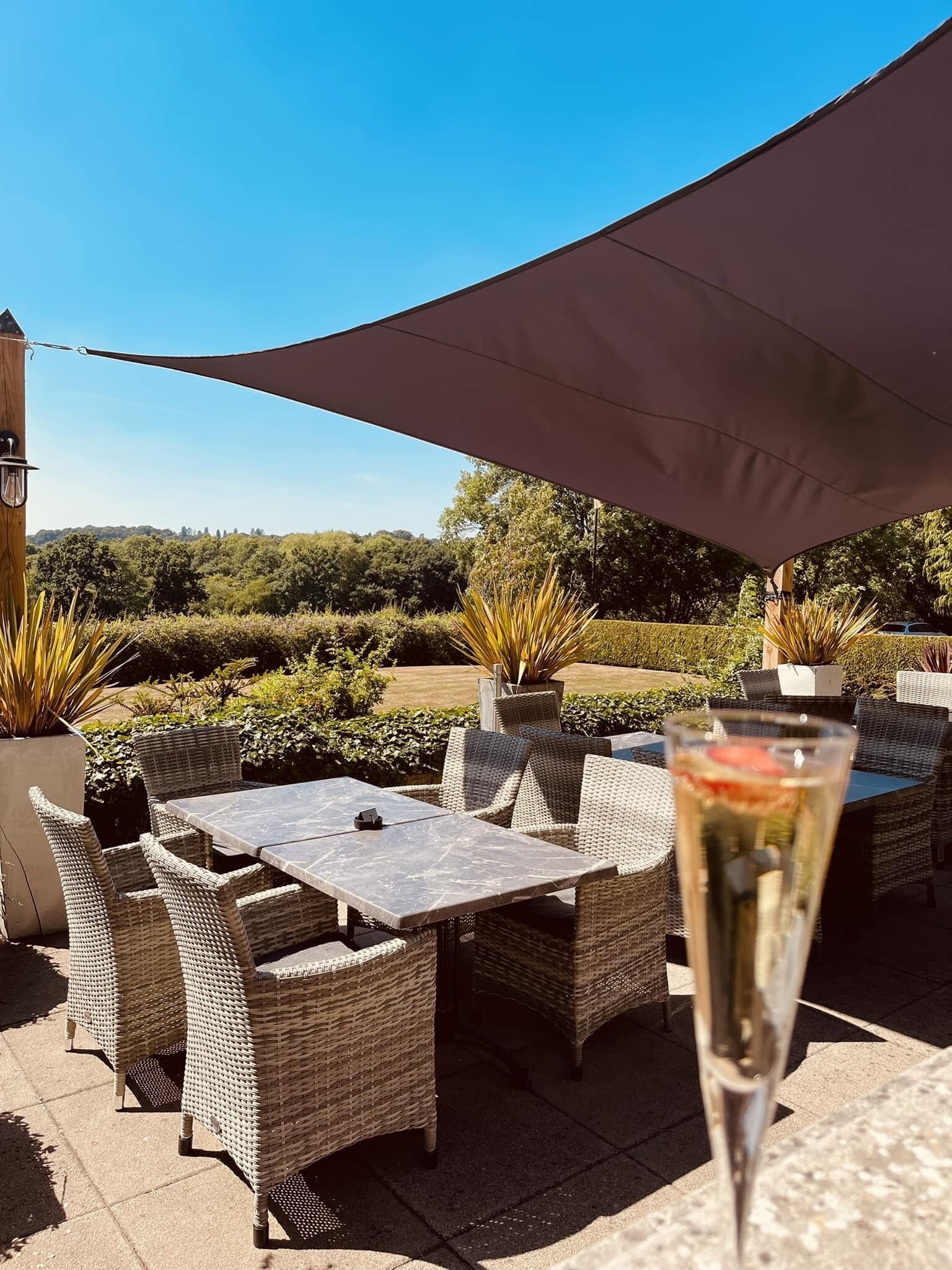 Une terrasse extérieure avec des meubles en osier et une table, un verre à portée de main, et une vue sur la verdure et les collines en arrière-plan.